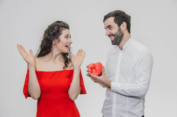 Young couple loving. A man made a present for his wife. During this time, the red field is presented in the form of a heart. Wife is pleasantly surprised. Dressed in a red dress and a white shirt. - Fotografie, Obrázek