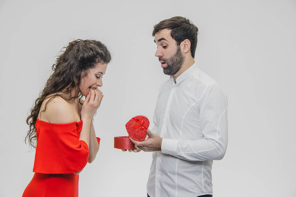 Young couple loving. A man made a present for his wife. During this time, the red field is presented in the form of a heart. Wife is pleasantly surprised. Dressed in a red dress and a white shirt. - Photo, Image