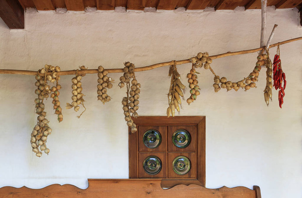 View of the terrace of an old Ukrainian rural hut with an antique window and pigtails of onion, corn and pepper drying on a wooden pole. - Zdjęcie, obraz