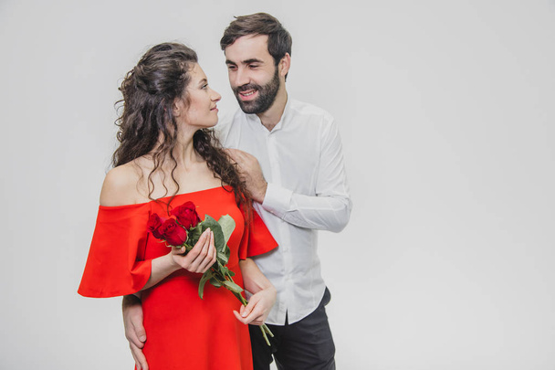 Elegant man gives a beautiful woman a red dress and a bouquet of red roses. Loving couple on a white background. - Fotografie, Obrázek