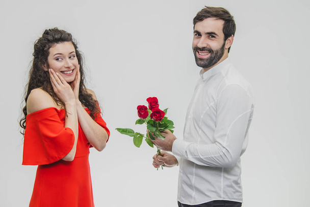 Elegant man gives a beautiful woman a red dress and a bouquet of red roses. Loving couple on a white background. - Φωτογραφία, εικόνα