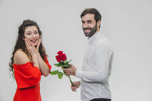 A young man hiding a bunch of roses behind his surprise to astonish his girlfriend for Valentines Day. Dressed in red dress and white shirt. - Zdjęcie, obraz