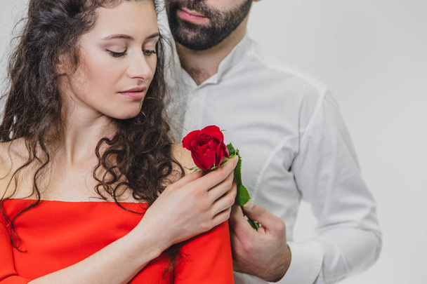 A young, handsome man gently covers his beautiful wife. During this time, one red rose. The girl is dressed in a red dress, a guy in a white shirt. On a white background. - Fotografie, Obrázek