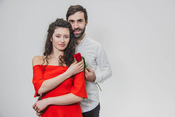 A young, handsome man gently covers his beautiful wife. During this time, one red rose. The girl is dressed in a red dress, a guy in a white shirt. On a white background. - Foto, Bild