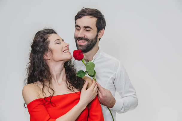 A young, handsome man gently covers his beautiful wife. During this time, one red rose. The girl is dressed in a red dress, a guy in a white shirt. On a white background. - 写真・画像