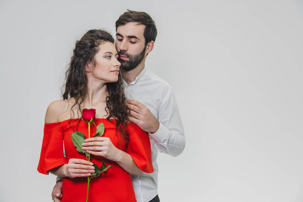 A young, handsome man gently covers his beautiful wife. During this time, one red rose. The girl is dressed in a red dress, a guy in a white shirt. - Foto, immagini