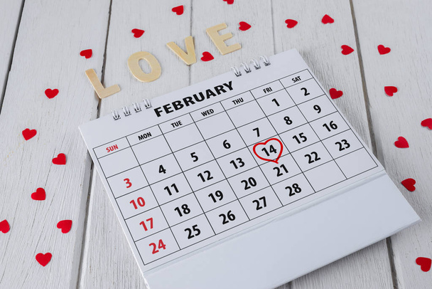 Calendar page with red hand written heart highlight on February 14 of Saint Valentines day with red Heart shape, Wooden letters word "LOVE" on white wooden table - Photo, Image