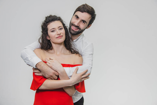 A young, sympathetic man gently embraces his beautiful wife. During this girl dressed in a red dress, a guy in a white shirt. On a white background. - Fotoğraf, Görsel