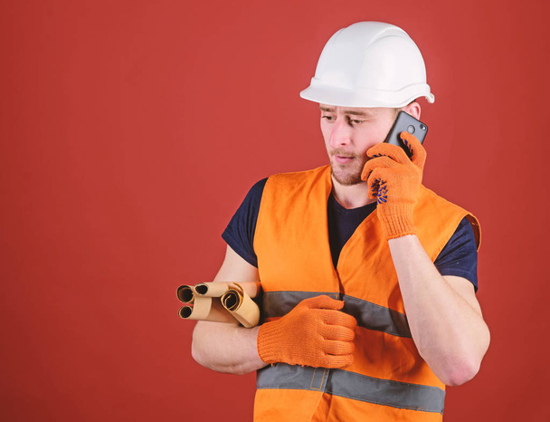Engineer, architect, builder on busy face speaks on smartphone while holds blueprints. Man, foreman in helmet supervises construction on phone, red background. Architect concept - Фото, зображення