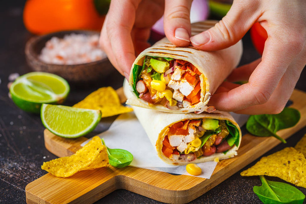 Grilled burritos wraps with chicken, beans, corn, tomatoes and avocado on a wooden board, dark background. Meat burrito, mexican food. - Photo, image