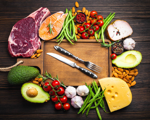 Knife and fork over wooden cutting board and ketogenic low carbs ingredients for healthy eating concept and weight loss, top view. Keto foods: meat, fish, avocado, cheese, vegetables, nuts - 写真・画像