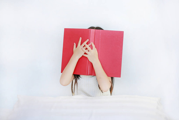 Little Asian child girl hiding face with open hardcover book sitting on bed with pillow. - Photo, image
