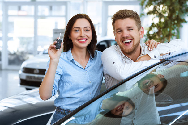 Наша первая машина. Счастливая женщина улыбается, показывая ключи от новой машины, которую они только что купили с мужем, счастливая пара улыбается фотоаппарату в дилерском центре
 - Фото, изображение