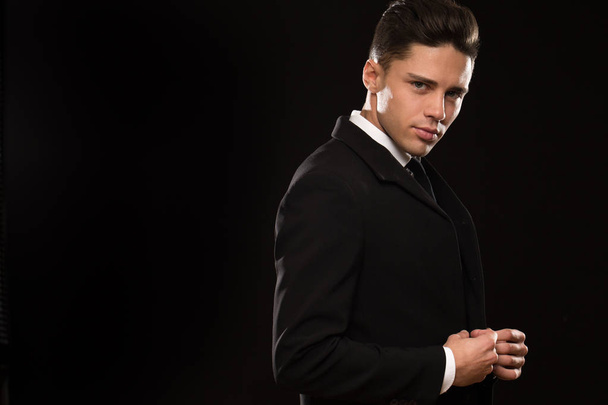Handsome sexy young man in a classy elegant black suit looking to the camera adjusting his jacket on black background copyspace sexuality seduction macho elegance businessman style - Fotó, kép