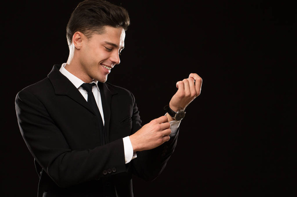 Wesoły przystojny młody człowiek uśmiechnięty z radością dostosowując rękawy jego koszula ubrana elegancka klasyczna czarna garnitur i krawat na czarnym tle sexy uwodzicielski sukces męskości - Zdjęcie, obraz