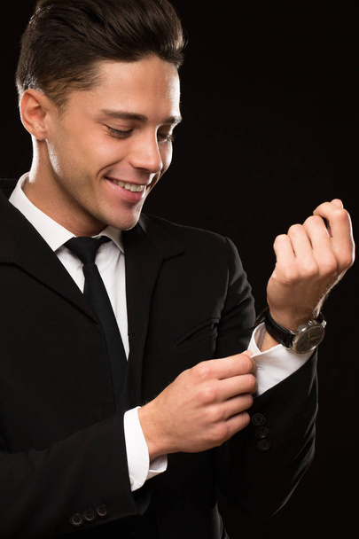 Vertikal ausgeschnittene Aufnahme eines gutaussehenden jungen Mannes, der freudig lächelnd die Ärmel seines Hemdes auf schwarzem Hintergrund anpasst, Macho-Positivität und Erfolgssicherheit im Stil von Geschäftsleuten - Foto, Bild