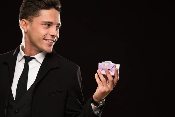 Cropped close up of a handsome young man in a black suit smiling happily holding a present looking at the copyspace celebration festive romance love relationships surprise dating gift flirt - Fotoğraf, Görsel