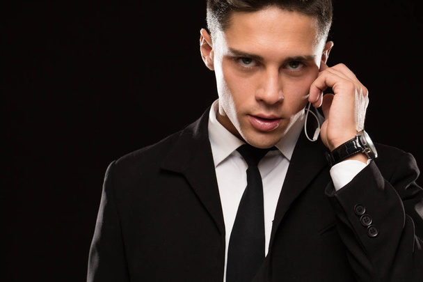 Close up of a handsome young suited secret service agent looking to the camera fiercely talking on his earphones black background copyspace infrmation security guarding bodyguard FBI secrecy - Photo, Image