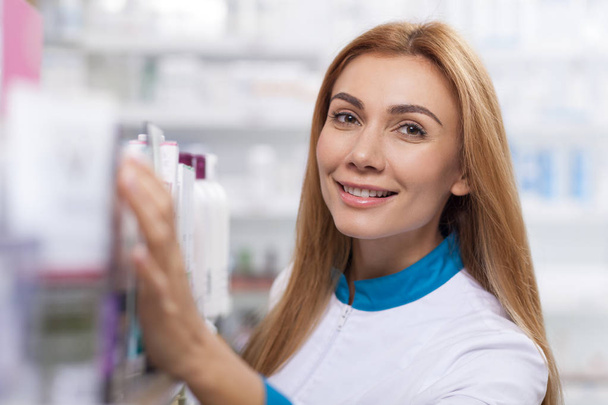 Close up of a cheerful drugstore worker smiling to the camera, while checking stock. Happy beautiful female pharmacist enjoying working at her pharmacy. Service, helpful staff, health concept - Photo, Image