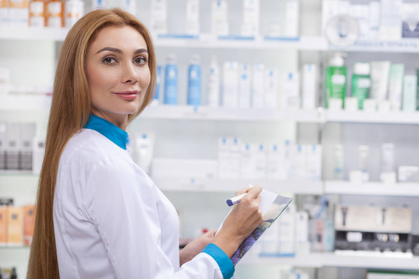 Happy professional chemist smiling to the camera, fillinf papers, working at the drugstore. Beautiful female pharmacist enjoying working at pharmacy. Insurance, patients support concept - Photo, Image