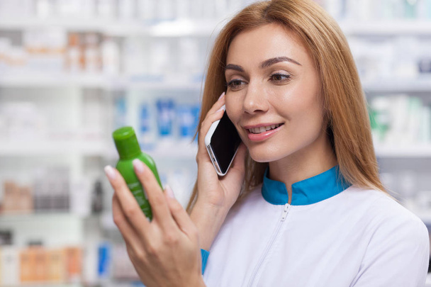 Friendly female pharmacist receiving orders via telephone, working at the drugstore. Cheerful chemist calling her patients, advising on medications. Communication, service, online shopping concept - Foto, Imagen