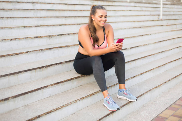 Young cheerful plus size woman in sporty top and leggings sitting on stairs happily using cellphone spending time outdoor - Foto, afbeelding