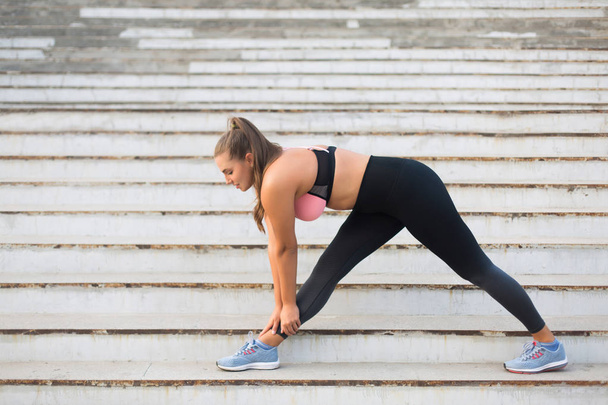 Young attractive plus size woman in sporty top and leggings stretching on stairs spending time outdoor - Photo, image