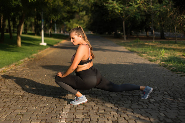 Jonge plus size vrouw in sportieve top en legging zorgvuldig doen sport oefeningen tijd doorbrengen in het gezellige stadspark  - Foto, afbeelding