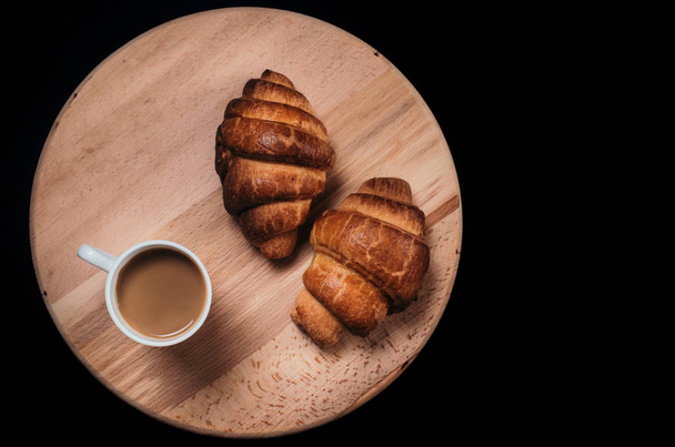 From above view of two fresh croissants and  mug with coffee placed on napkin on black background of table - Foto, afbeelding
