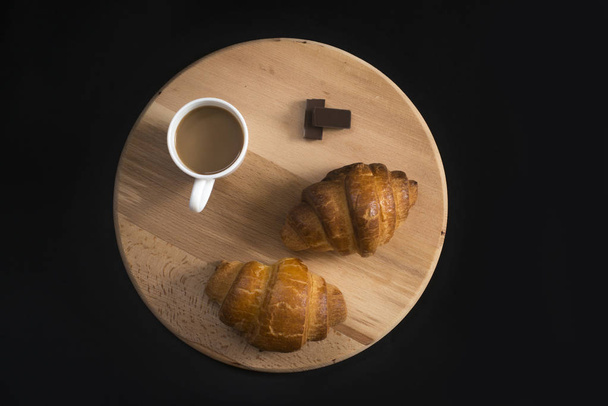 From above view of two fresh croissants, pieces of chocolate and  mug with coffee placed on napkin on black background of table - Fotó, kép