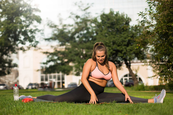 Młody zmęczony plus rozmiar Kobieta w różowy top sportowy i legginsy próbuje siedzieć na sznurek na matę do jogi spędzać czas w parku miejskim  - Zdjęcie, obraz