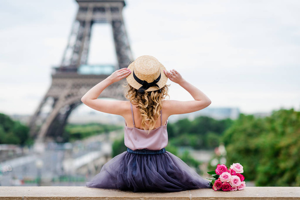 Blond girl with curls holding her hat and looking at the Eiffel tower in Paris from the roof top of the Trocadero place - Photo, Image