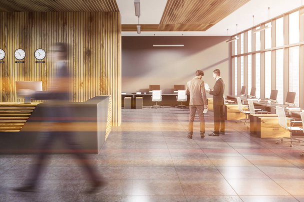 Businessmen in interior of modern office with gray and wooden walls, tiled floor, rows of computer tables with white chairs and gray and wooden reception desk. Toned image blur - Foto, afbeelding