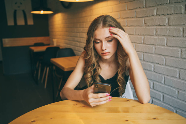 Arayan bir kafede oturan üzgün genç kız yorgun onun telefon, siyah telefonunu izlerken mutsuz kız, - Fotoğraf, Görsel