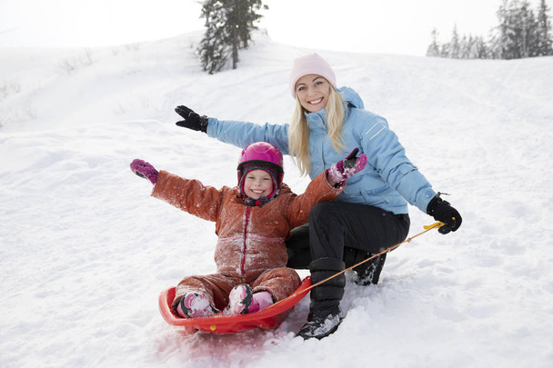 Mom and daughter ride on a sled from a snow slide. Ride from a snow hill on a sled. Sleigh rides, winter fun, snow, family sleigh rides. - 写真・画像