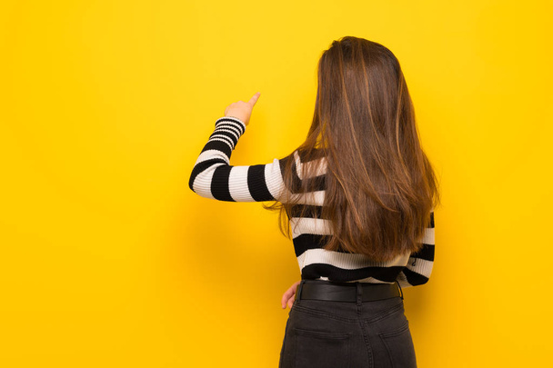 Jovem mulher sobre a parede amarela apontando para trás com o dedo indicador
 - Foto, Imagem