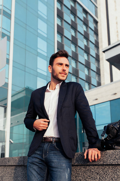 handsome young businessman with a beard and in a business suit standing on the street against the background of the office building next to a comfortable stylish leather bag. - 写真・画像