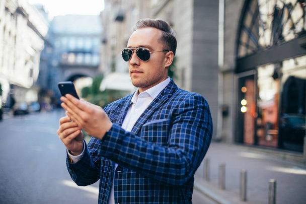 Πορτρέτο του κομψό όμορφος νεαρός άνδρας στέκεται σε εξωτερικούς χώρους. Άντρας με σακάκι και πουκάμισο. άνθρωπος που χρησιμοποιεί κινητό τηλέφωνο - Φωτογραφία, εικόνα