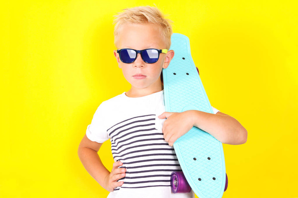 helle Portpait auf gelbem Hintergrund von niedlichen coolen blonden Jungen trägt Sonnenbrille mit Skateboard in den Armen - Foto, Bild