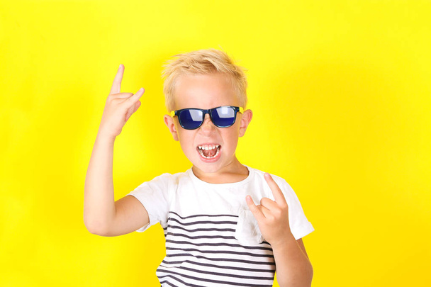 Schattige blonde jongen dragen van een zonnebril op gele achtergrond met hoorns gebaar - Foto, afbeelding