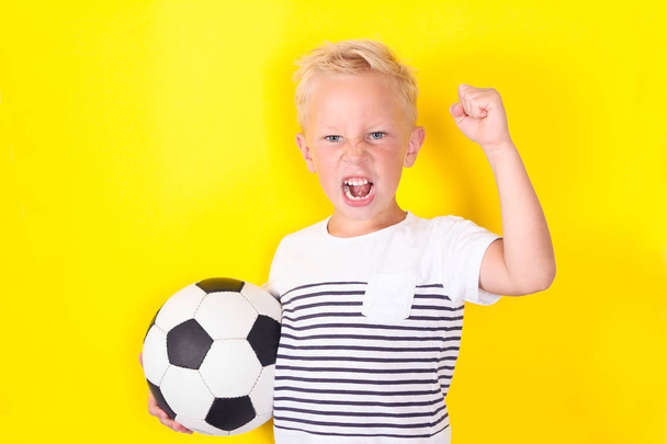 Cute blond boy portrait with ball on yellow background locking football game - Foto, afbeelding