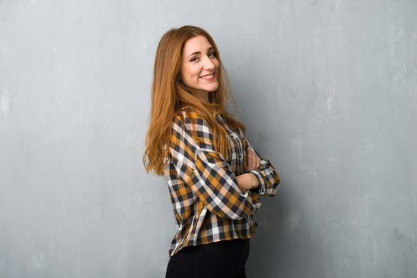 Young redhead girl over grunge wall keeping the arms crossed in lateral position while smiling - Φωτογραφία, εικόνα