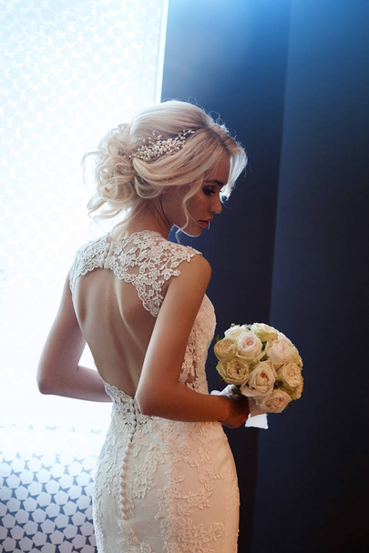 Morning bride. A woman in a white wedding dress holding a bouquet of flowers in her hands. Beautiful blonde girl getting ready for wedding ceremony - Foto, Imagem