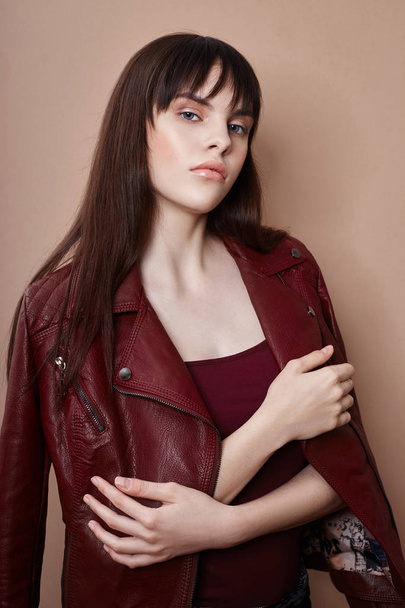 Sexy brunette in leather jacket and red top, portrait bright makeup and lipstick close-up, face makeup, professional makeup, skin care. Young sexy woman posing on beige background, flying hair - Fotó, kép