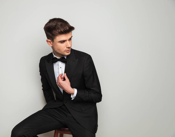 curious groom holding tuxedo collar looks to side while sitting on wooden chair - 写真・画像