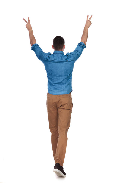 rear view of celebrating casual man walking on white background and making peace sign, full length picture - Photo, Image