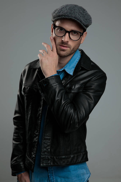 portrait of seductive fashion man in leather jacket touching his face while standing on light grey background with hand in pocket - Photo, Image