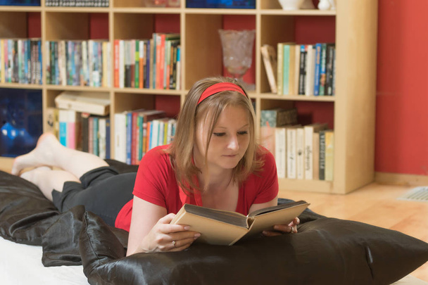 Cool attractive young woman in red is reading a book lying on the floor indoors.  - Valokuva, kuva