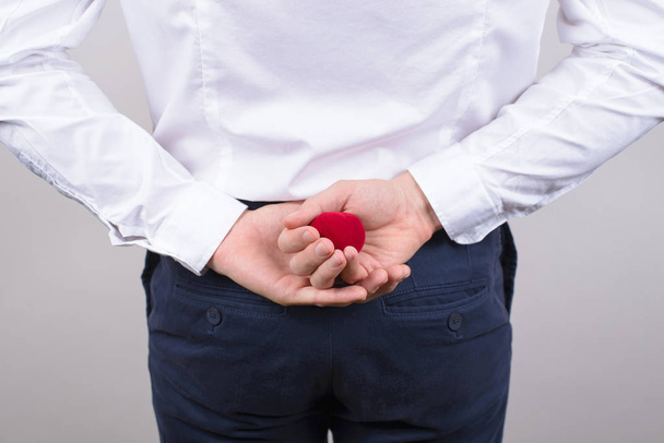 Беддинд вид сзади фото взволнованного нервного джентльмена, прячущего небольшой ярко-красный ящик с кольцом за спиной изолированный серый фон
 - Фото, изображение