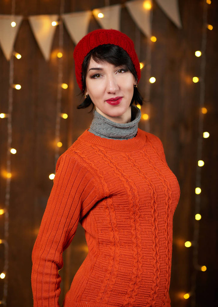 Hermosa mujer está posando sobre fondo de madera oscura, luces y banderas de Navidad, concepto de vacaciones. Vestido con sombrero y suéter de punto rojo
. - Foto, imagen